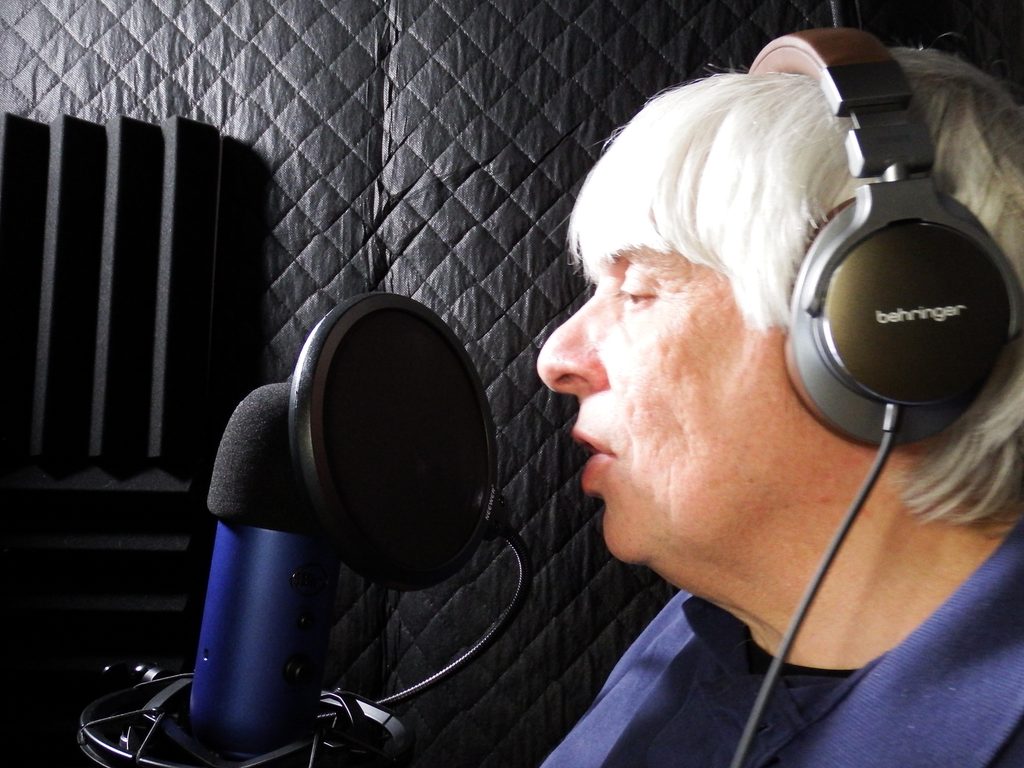 Jimmy in the studio recording at Jimmy's Voice Over studio. Jimmy with headphones, talking into a pro Blue Yeti microphone with black sound blankets and black sound proof foam panels.
